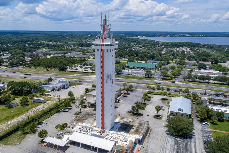 Aerial view over a Central Florida town with a tall observation tower beside the highway corridor, tree-shaded neighborhoods and a lake beyond, representative of the Clermont area