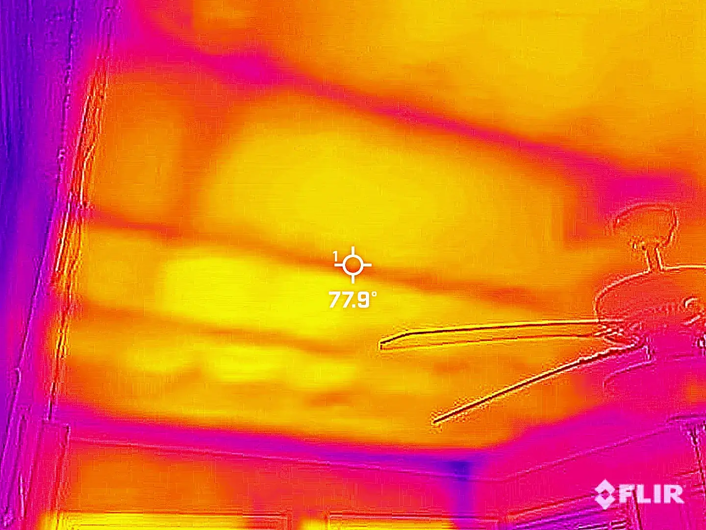 Infrared thermal image of a ceiling in which the framing above reads as cooler bands against the warmer ceiling surface, with an on-screen crosshair reporting a spot surface temperature.